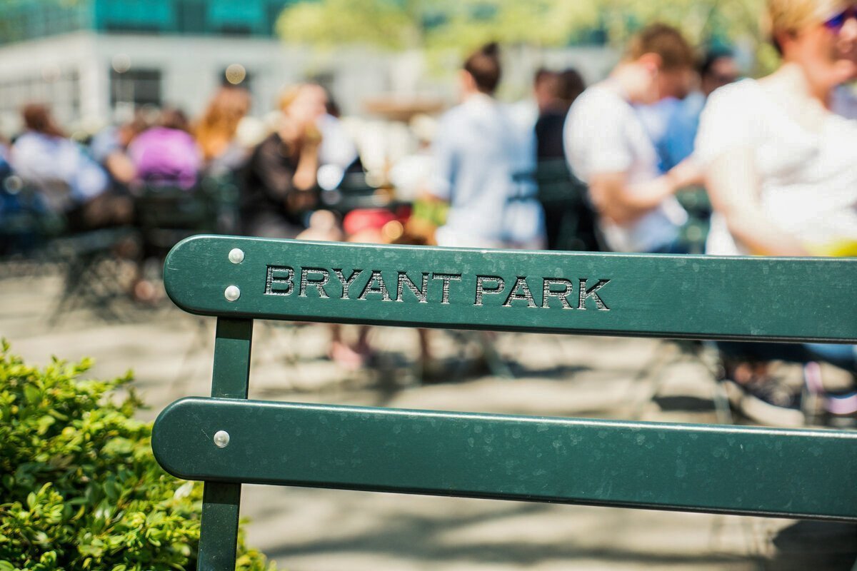 Bryant Park name embossed on a chair with tourist and New Yorkers enjoying lunchtime in the background.