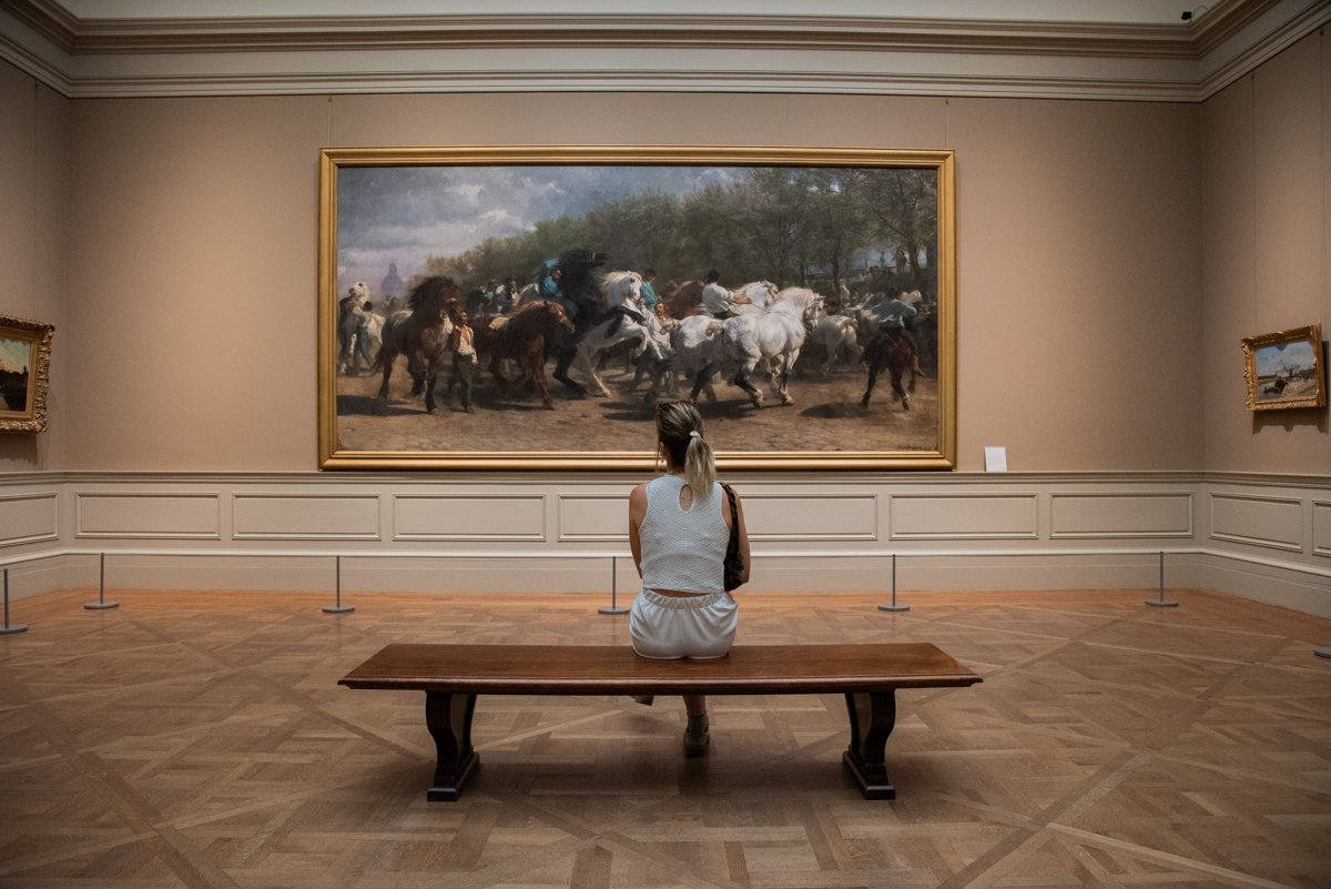 Femme assise qui regarde une grande peinture de cavaliers et chevaux dans une salle
